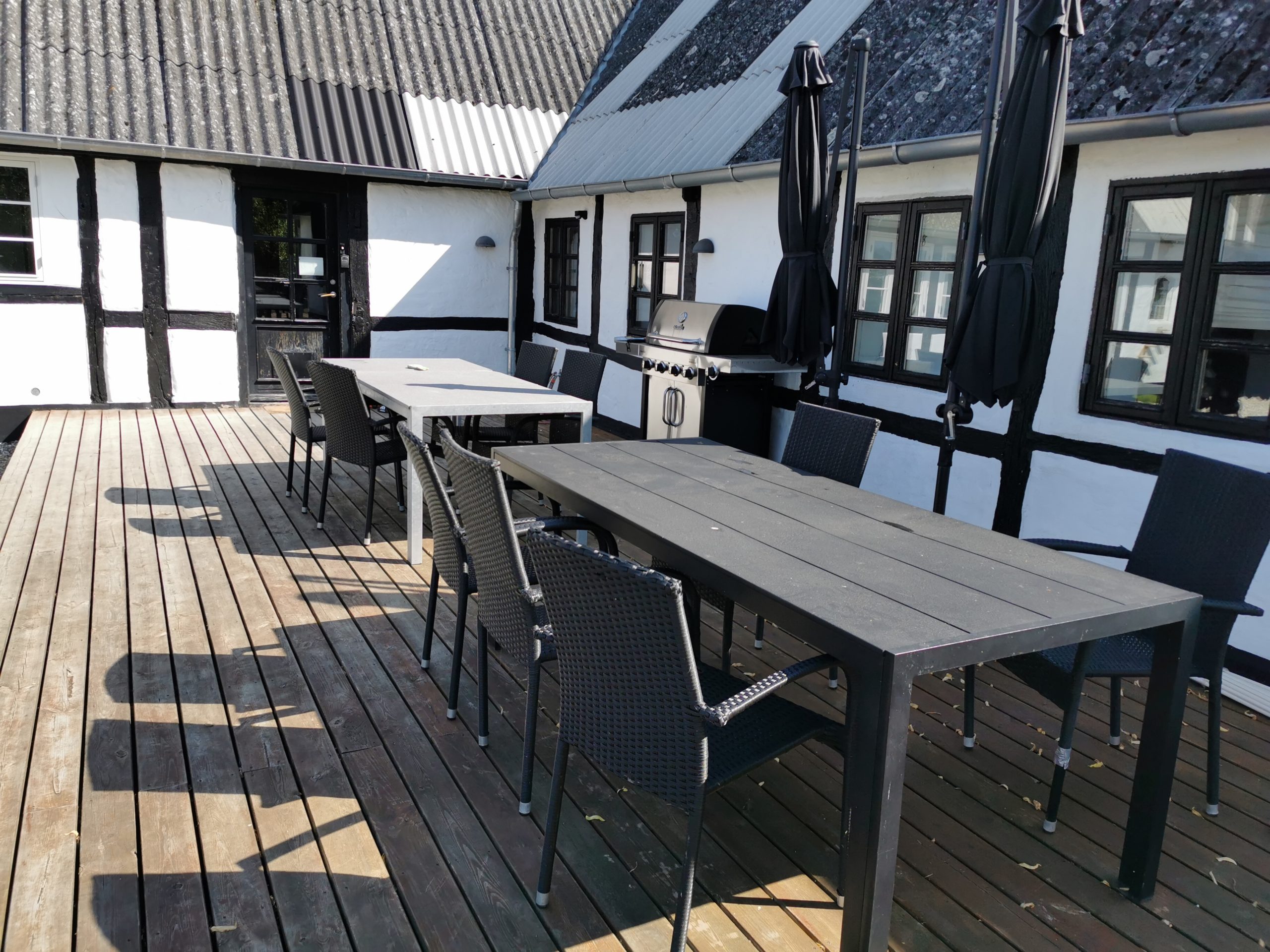 Udendørs træterrasse med to lange sorte haveborde og flere sorte fletstole, sammenfoldede sorte parasoller og en gasgrill opstillet mod et hvidt bindingsværkshus med sorte bjælker; solskygger falder henover træbrædderne.