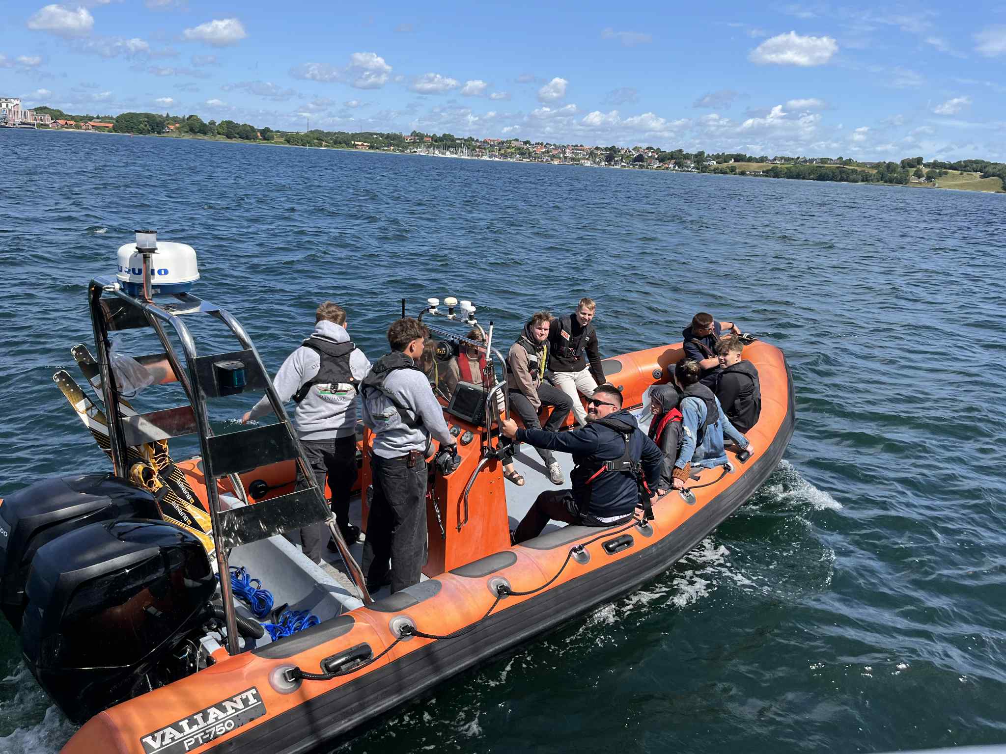 Orange RIB-båd mærket u0022VALIANT DT-750u0022 med otte-ni personer, nogle siddende og andre stående iført redningsveste, sejler på mørkeblå havvand tæt på en kystlinje under en solskinsdag.