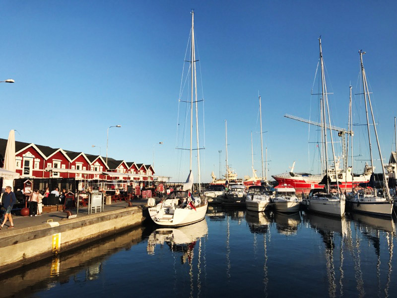 Sejlbåde fortøjet i en marina, deres master spejles i det stille vand. Røde kajhuse og caféer langs venstre kaj med gående mennesker, samt en havnekran og større skibe i baggrunden under en klar blå himmel.