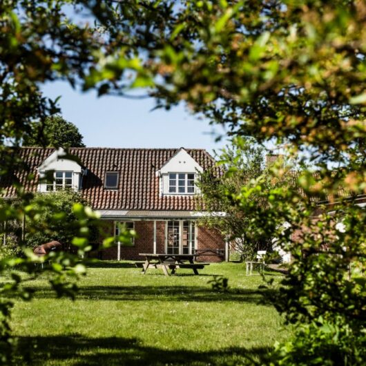Rødt teglhus med hvide kvistvinduer og rødt tegltag set gennem en ramme af grønne buske og træer. Foran huset en solbeskinnet græsplæne med en træ-picnicbænk og en enkelt hvid stol.