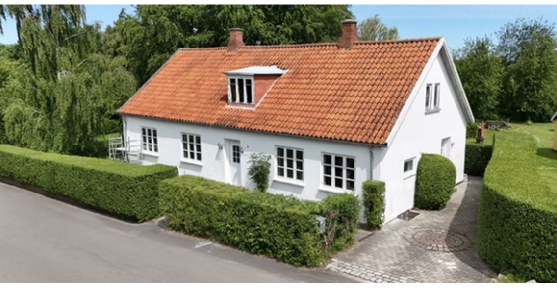 Hvidt parcelhus med orange tegltag og en lille kvist på taget set skråt forfra; huset er omgivet af velklippede hække langs vejen og har en brostensindkørsel til højre med træer i baggrunden.