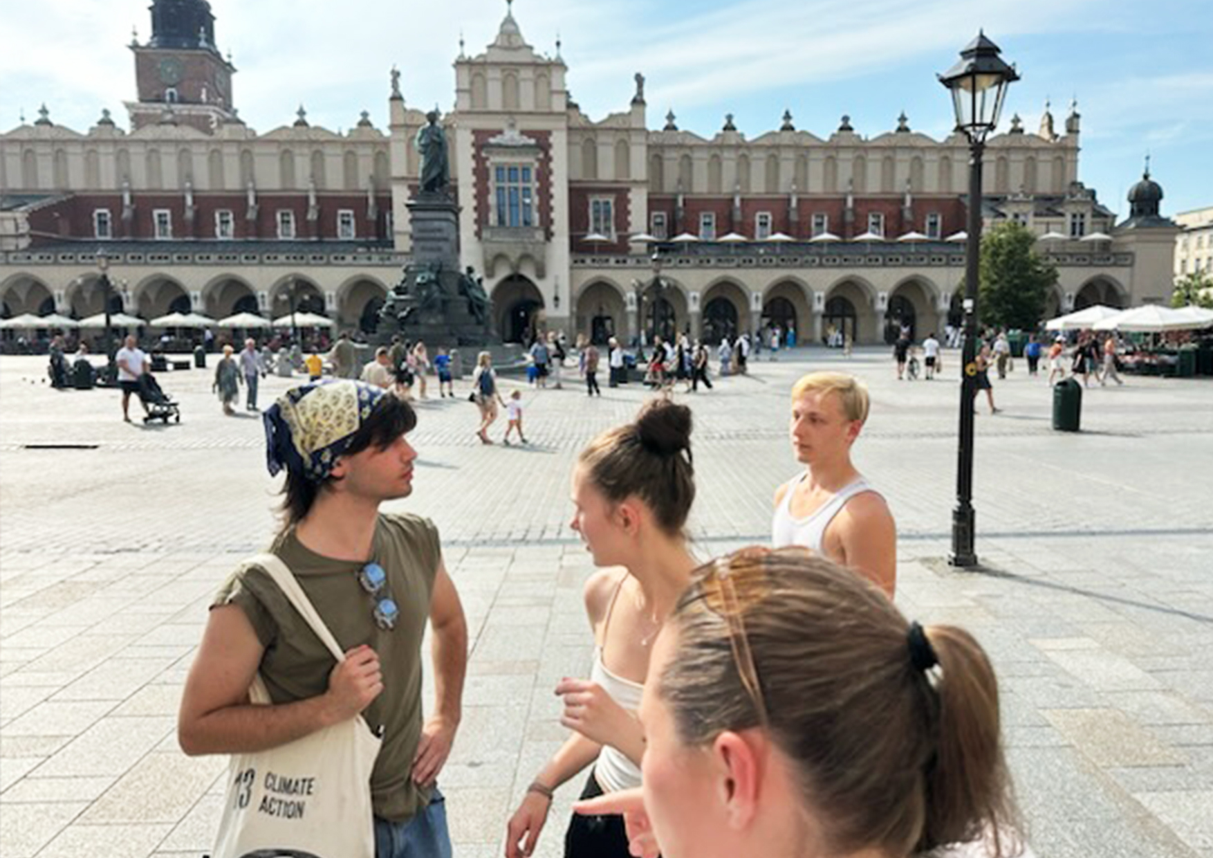 Fire unge personer står og taler på en brostensbelagt plads foran en stor historisk bygning med buegange og en statue i baggrunden; på en af de unges stoftaske står teksten u002213 CLIMATE ACTIONu0022.