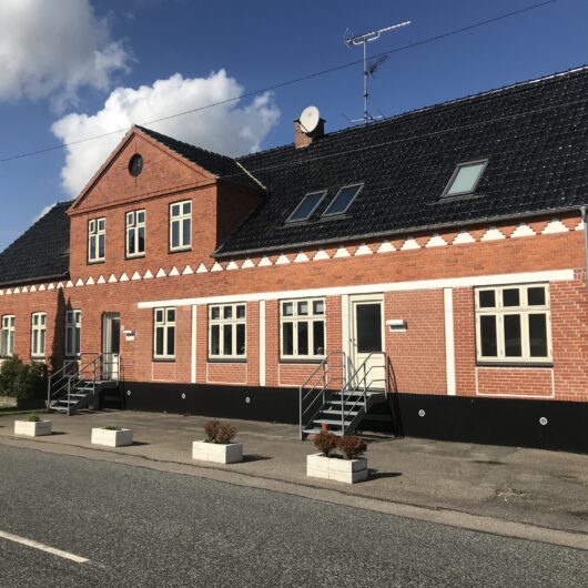 Rødt to-etagers murstenshus med sort saddeltag og hvide vinduesrammer set fra fortovet; to metaltrapper fører op til indgangsdøre, og satellitantenner sidder på taget. Blå himmel med få hvide skyer og fire hvide plantekasser står langs fortovets kant.