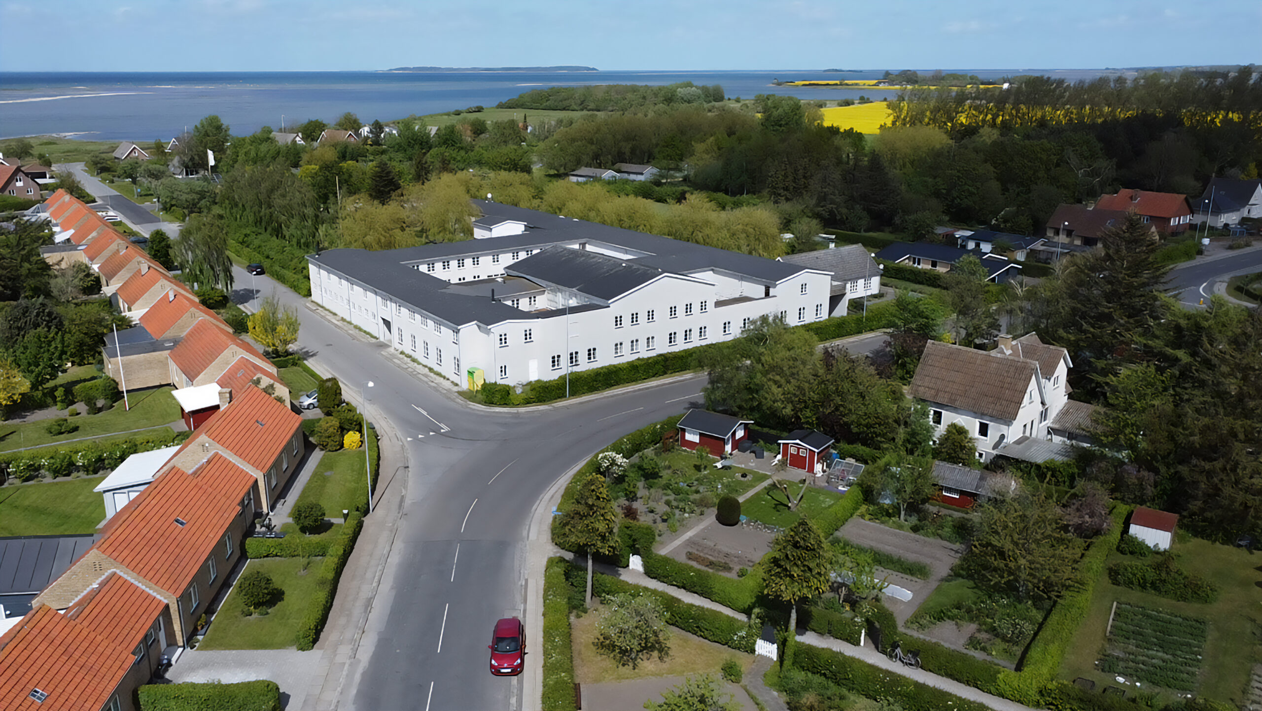 Luftfoto af en kystlandsby med en stor hvid U-formet bygning ved et vejkryds, omgivet af rækker af lave boliger med røde tegltage og velplejede haver. I forgrunden kører en rød bil på vej, og i baggrunden ses marker, skov og havkyst.