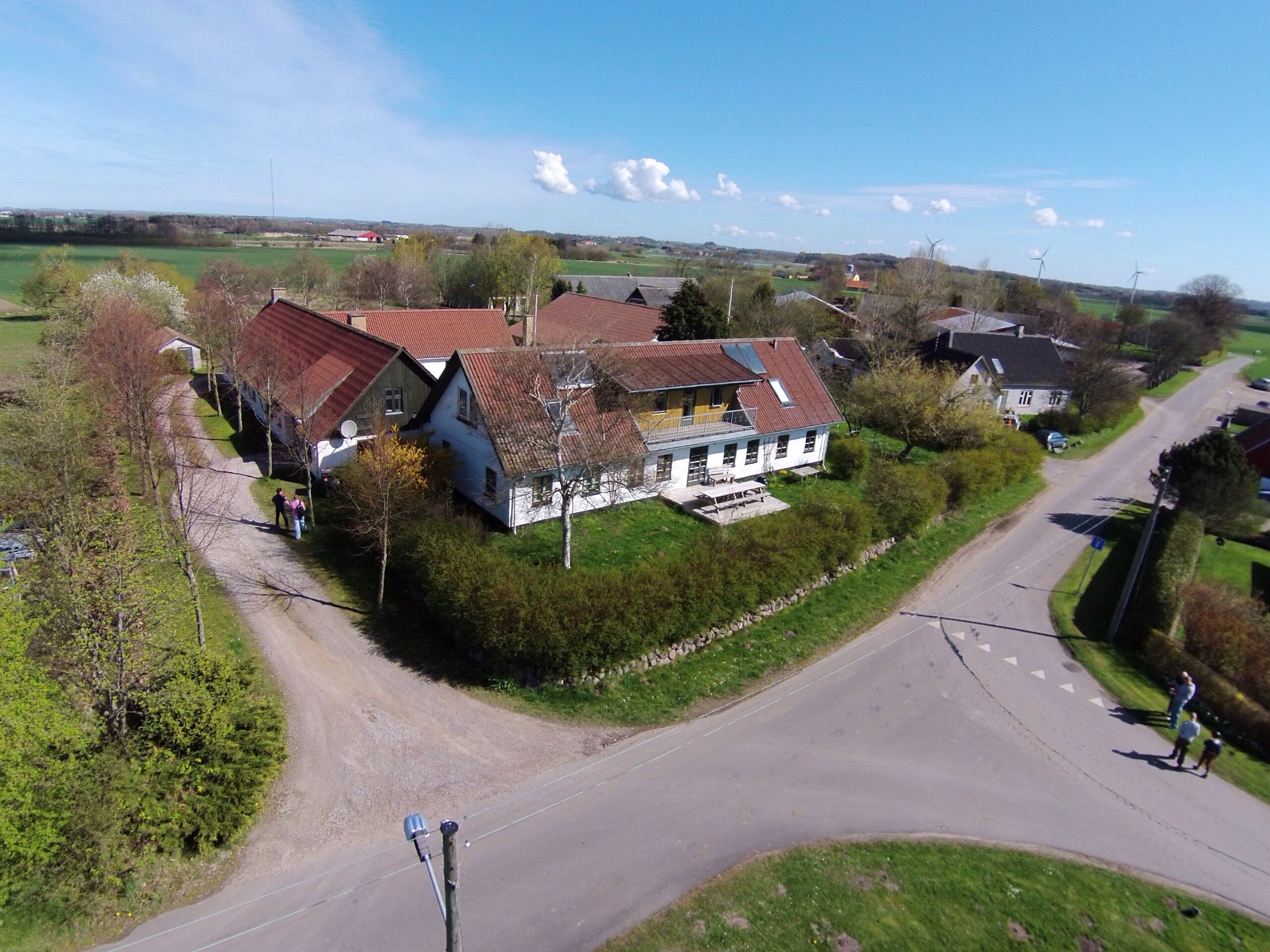 Luftfoto af en lille gårdgruppe med røde tage placeret ved et trekantet vejkryds i åbent land. Et par mennesker går på grusvejen tæt ved husene, omgivet af hække, marker og vindmøller i horisonten under en klar blå himmel.