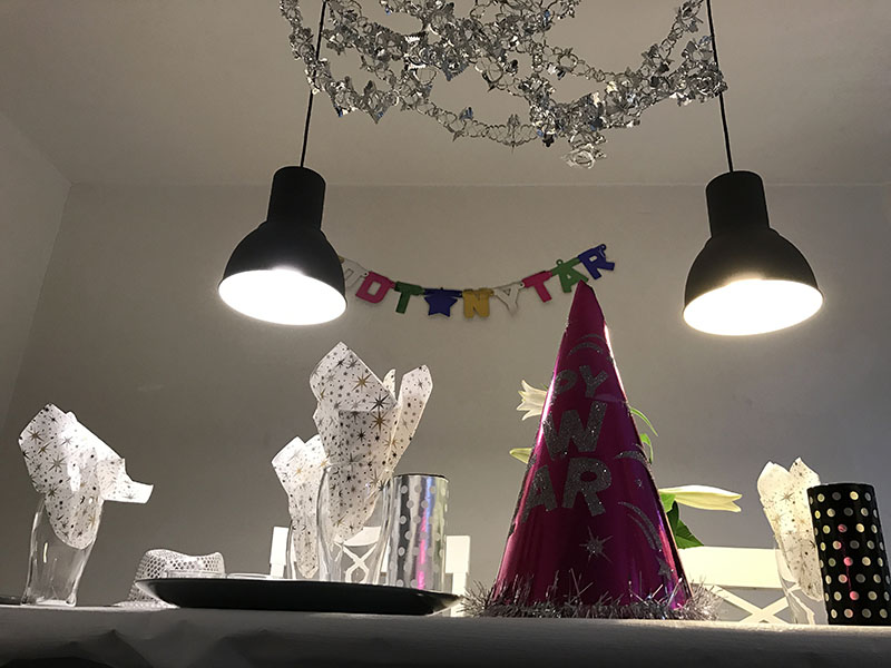 Bord pyntet til nytår under to hængende lamper med en lyserød konisk festhat i forgrunden med sølvtekst u0022HAPPY NEW YEARu0022 og en farverig guirlande på væggen med teksten u0022GODT NYTÅRu0022.
