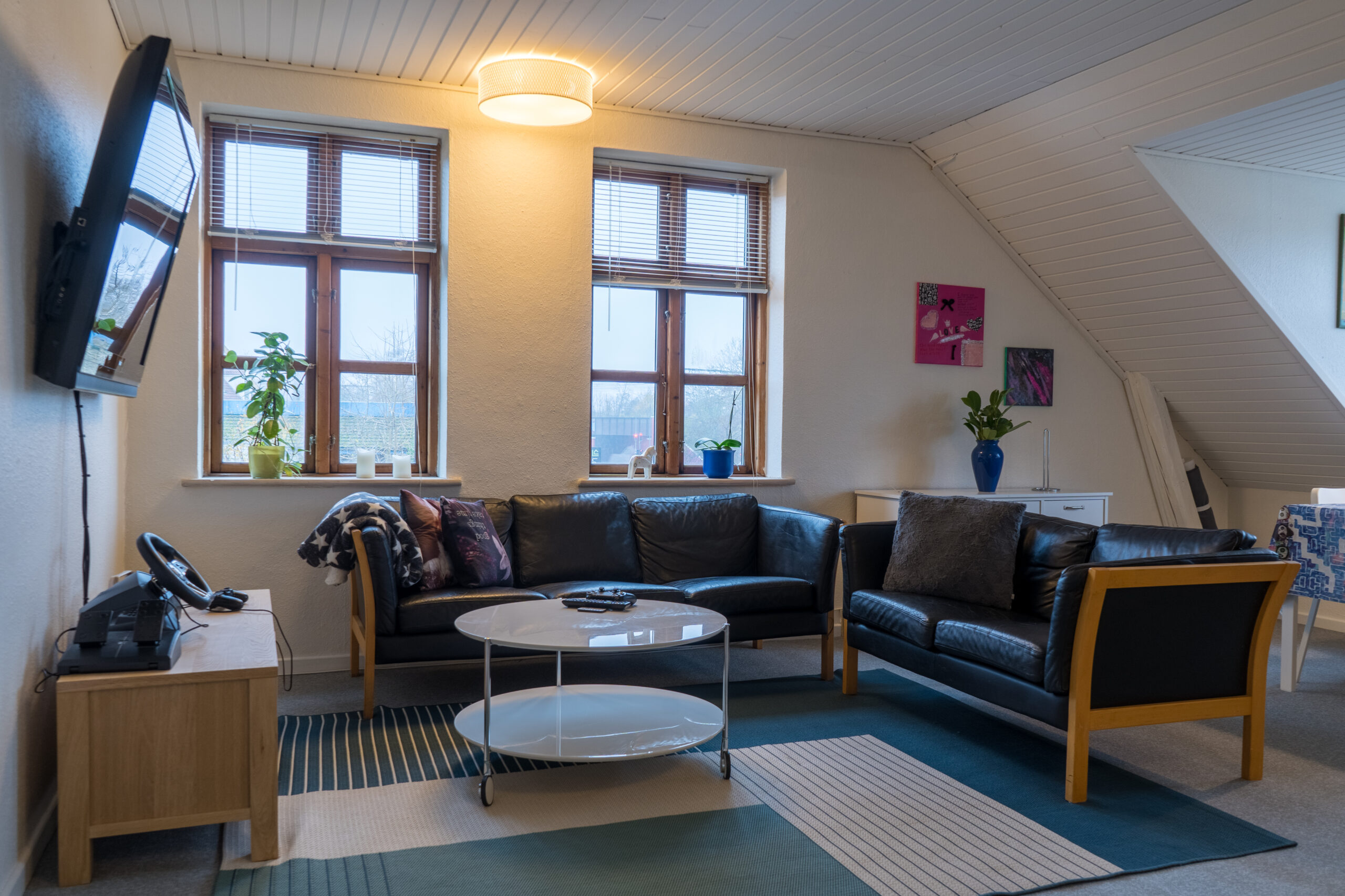 Stue med sort lædersofa i L-form og en matchende sofa med træramme omkring et rundt hvidt sofabord på et blåt og beige mønstret tæppe, med fjernbetjening på bordet. To vinduer med potteplanter under et skråt loft og et vægmonteret fjernsyn til venstre.