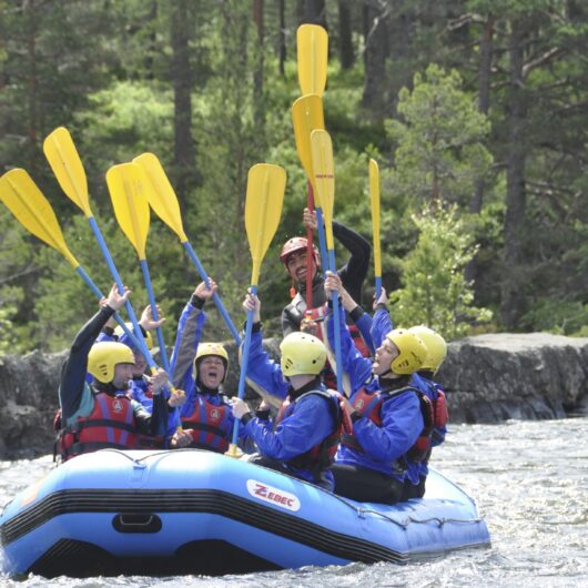 En gruppe raftere iført gule hjelme, blå jakker og røde redningsveste sidder i en blå oppustelig gummibåd og løfter deres gule årer i luften på en strømmende flod omgivet af klipper og skov; tekst "ZEBEC" ses på bådsiden.