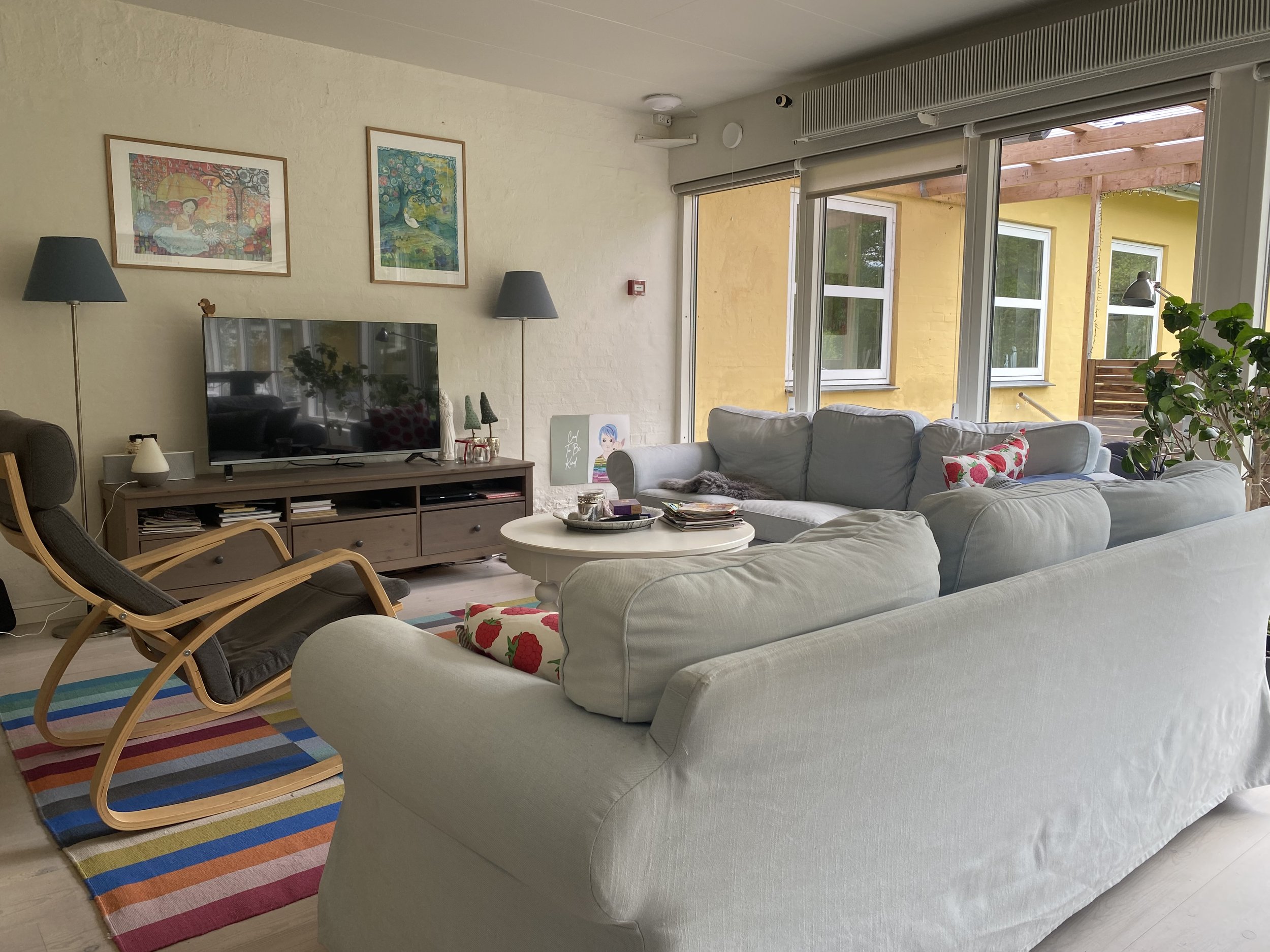 Stor lysegrå sofa i forgrunden samlet omkring et rundt hvidt sofabord i en lys stue; et fladt tv står på et lavt træskab foran, en buet træ-lænestol og to gulvlamper flankerer tv'et, og store skydedøre åbner ud til en gul facade.