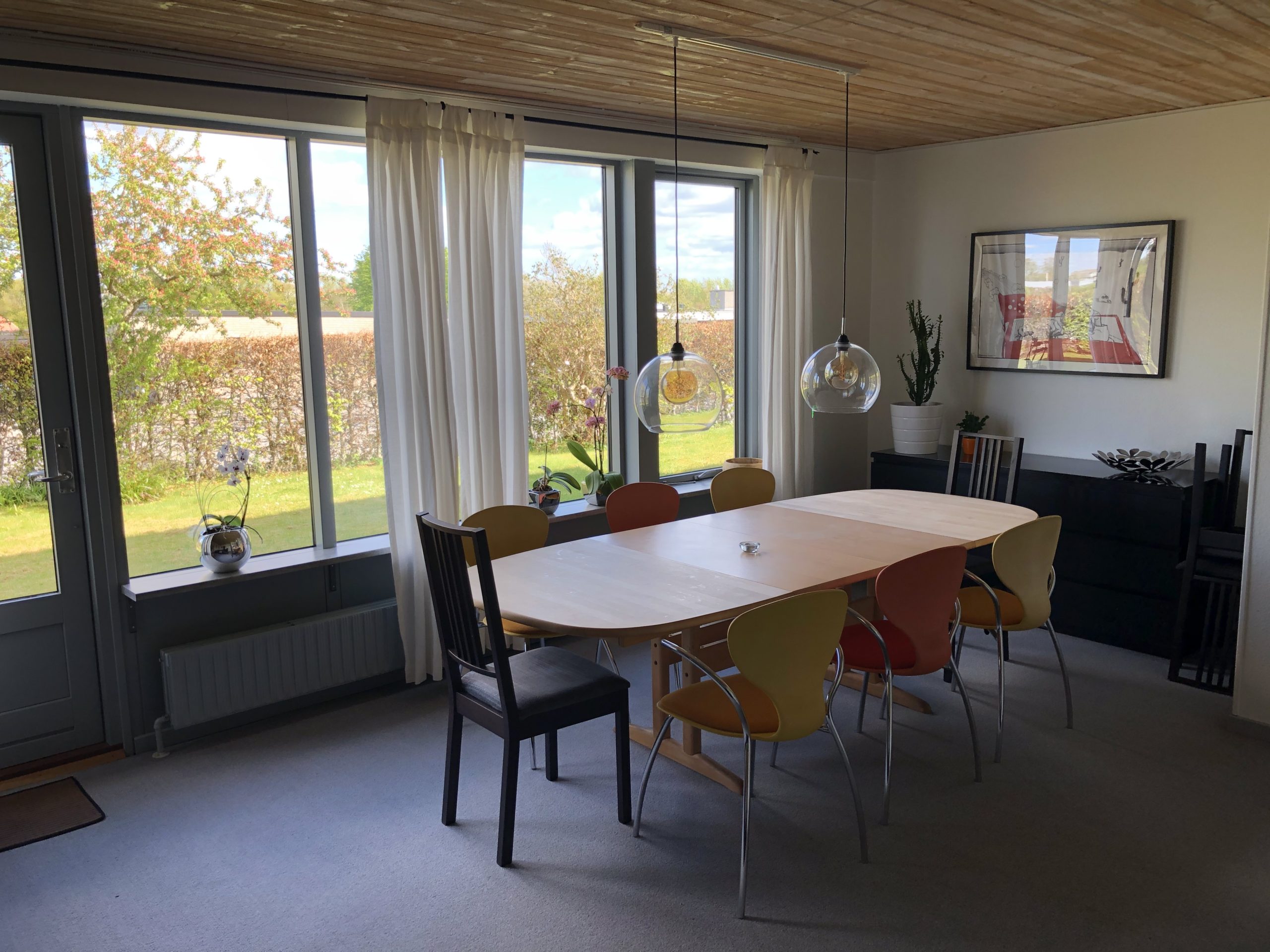 Lyst træspisebord omgivet af flere farvede stole og en sort stol i et lyst spisestue med store vinduer og hvide gardiner, to runde glaspendler hængende over bordet og udsigt til en have.