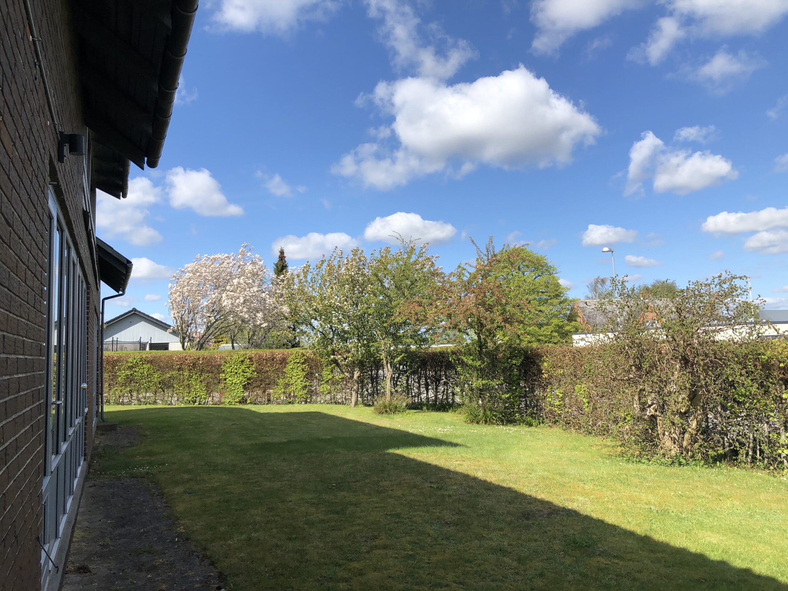 Murstensfacade til venstre med rækker af vinduer, der kaster en bred skygge over en grøn græsplæne; i baggrunden ses klippede hække og blomstrende træer under en klar blå himmel med spredte hvide skyer.
