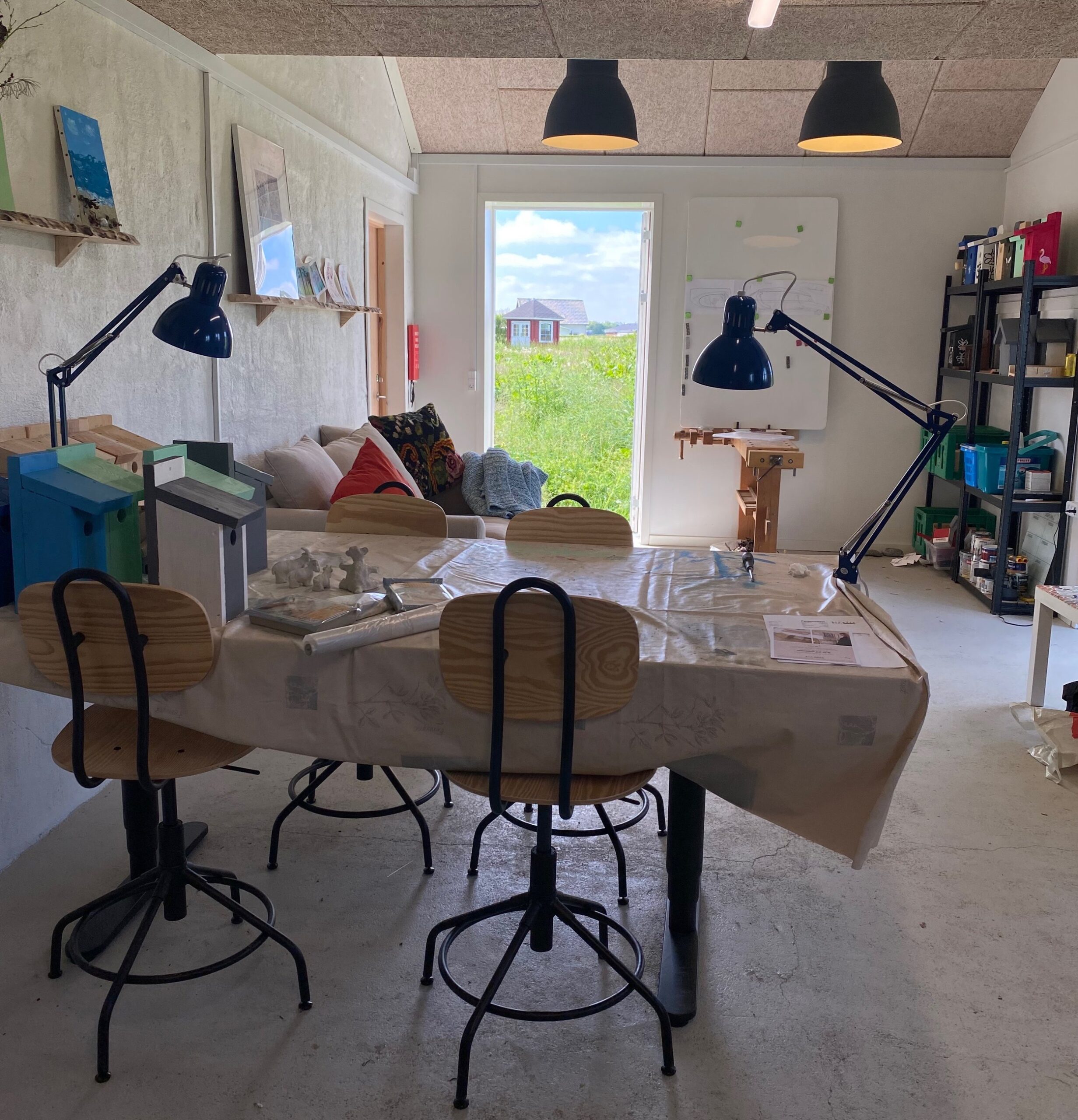 Et lyst værksted med et stort bord dækket af en plastikdug, fire træstole på sort metalstel og to store sorte arbejdslygter rettet mod bordet. I baggrunden ses en sofa, opbevaringshylder langs væggen og en åben dør ud til grønt græsland med en lille rød hytte under blå himmel.