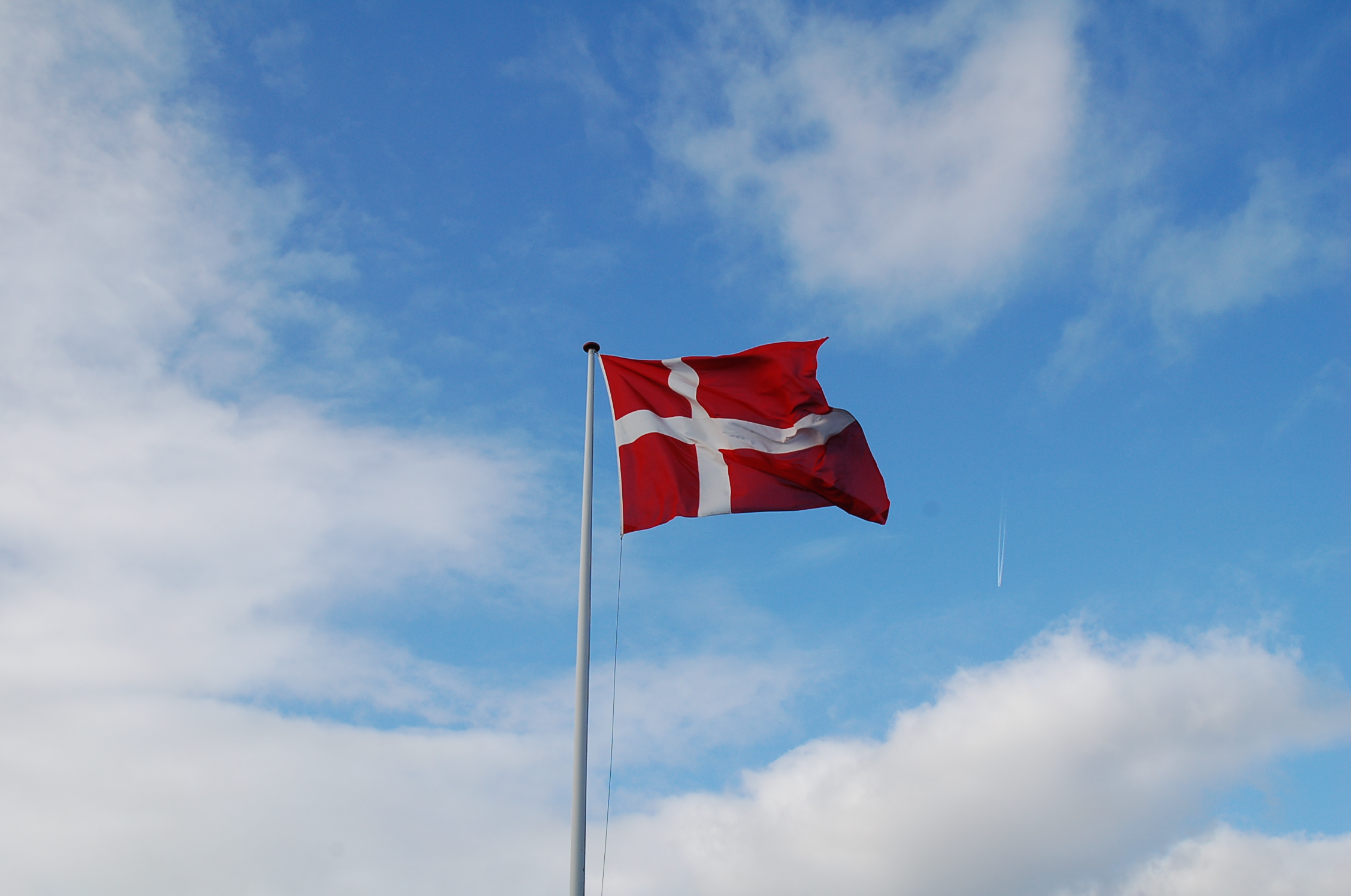Et rødt Dannebrog med hvidt kors blafrer på en høj flagstang mod en blå himmel med spredte hvide skyer og et svagt flyspor i baggrunden.