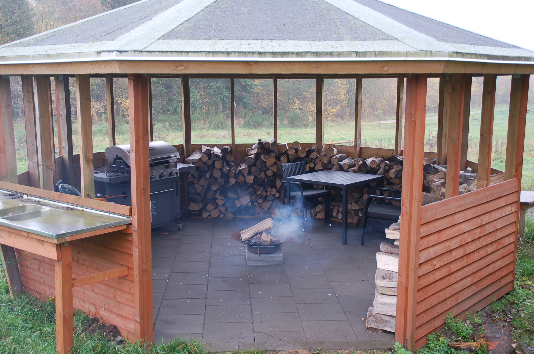 Træpavillon med åbne sider i et græsland, hvor en stor bunke brænde er stablet langs bagvæggen, en gasgrill står til venstre og et bord med stole til højre. Et lille bål i en metalskål ryger i midten af pavillonen, og en udendørs vask er monteret ved åbningen til venstre.