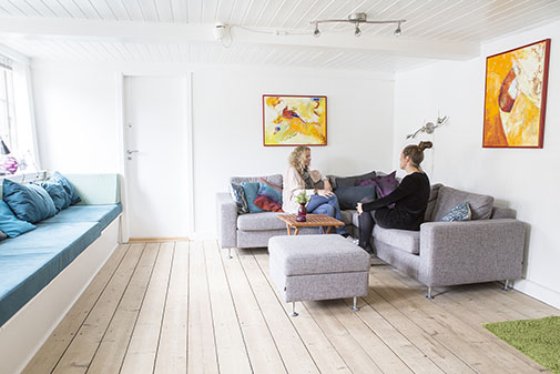 To kvinder sidder og samtaler på grå sofaer i en lys stue med et lille træbord og en grå skammel foran dem. Rummet har lyst trægulv, en vinduesplads med blå hynder til venstre og to farverige abstrakte malerier på hvide vægge.