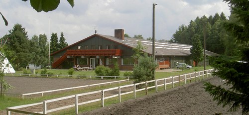 Lang, lav træbygning med skrånende tag, som ligger bag en sandet ridebane afgrænset af et hvidt træhegn; bygningen er omgivet af græs og nåletræer under en overskyet himmel.