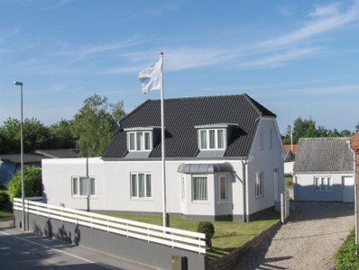 Et hvidt to-etagers hus med sort tag står ved en vej; en hvid flagstang i forhaven vajer med et hvidt flag, en lav hvid træhegn afgrænser græsplænen, og en grusindkørsel fører til en garage til højre.