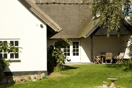 Hvidt sommerhus med stråtag og dobbelte franske døre i midten; vindue til venstre og buske langs husmuren. Grøn græsplæne i forgrunden, et fuglebad til højre, og et træbord med stole under en overdækket terrasse til højre.