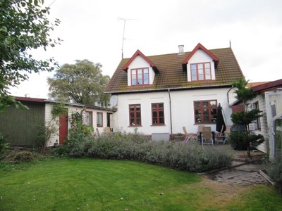 Et to-etagers hvidt hus med røde vinduesrammer og to kvistvinduer ses fra haven. Foran huset er en velplejet græsplæne, et lavt buskbed og en terrasse med havemøbler, flankeret af mindre udbygninger under en overskyet himmel.