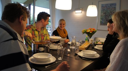 Fem personer sidder omkring et mørkt spisebord og deler et måltid; en person skænker mad fra en gryde midt på bordet, som er dækket med hvide skåle, glas, bestik, brød og en buket blomster under to hængelamper i en lys stue.