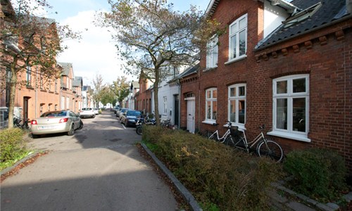 Række af røde murstensboliger langs en stille, træbevokset villavej med cykler lænet op ad husfacaderne og parkerede biler langs vejens kant på en lys dag.