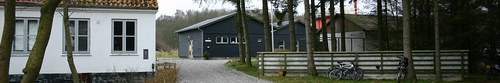 En grusvej fører fra forgrunden til en mørkegrå bygning midt i billedet, med et hvidt hus til venstre og et lavt træhegn til højre, hvor to cykler står lænet opad, omgivet af træer.