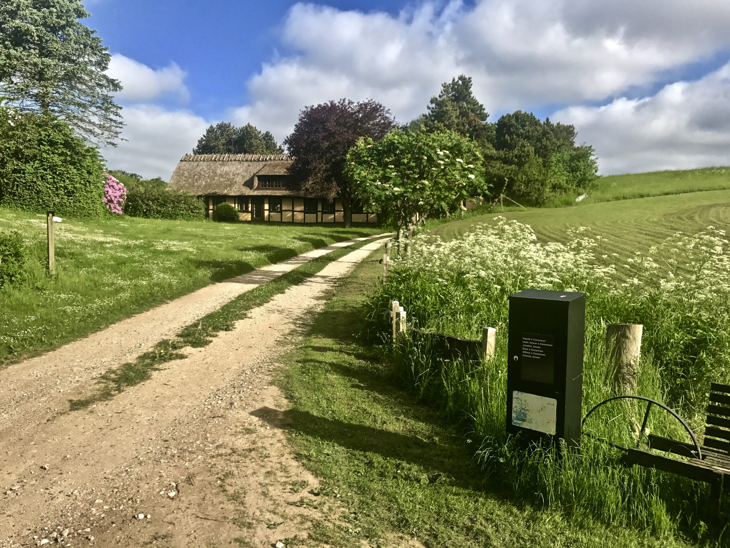 Grusvej fører forbi grønne marker og blomstrende grøftekant mod et stråtækt bindingsværkshus under en blå himmel med hvide skyer. Til højre i forgrunden står en sort informationsstander og en træbænk ved et lavt træhegn omgivet af højt græs.