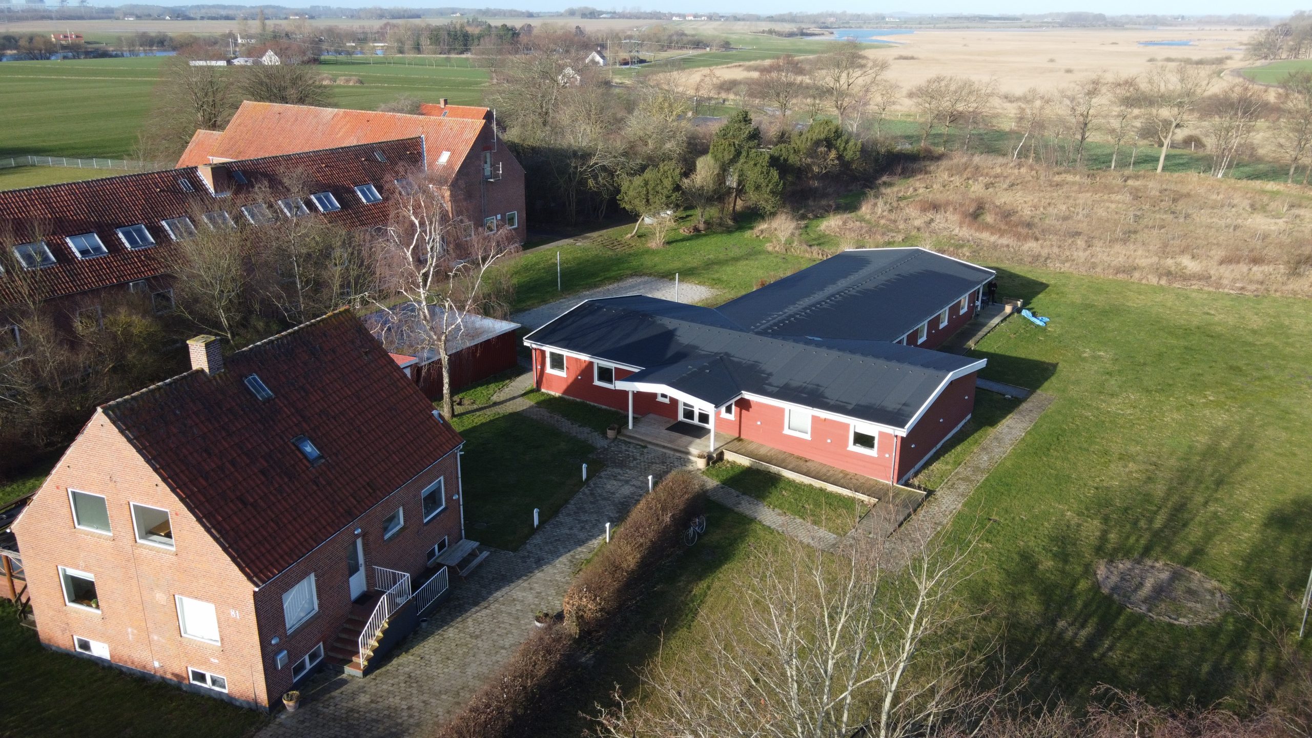 Luftfoto af to røde bygninger placeret på en stor græsplæne i et åbent landligt område; forrest et to-etagers murstenshus med rødt tegltag og indgangstrappe, bagved en lang, lav rødmalet bygning med sort fladt tag og omkringliggende stier og marker.