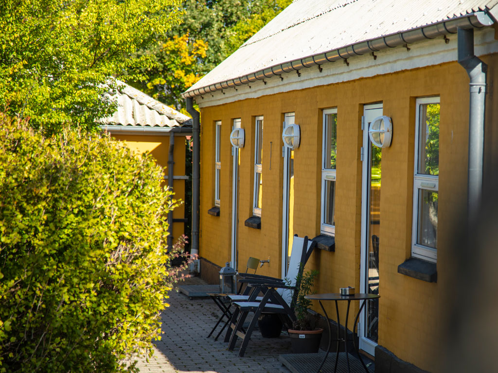 Række af gulmalede lejligheder med flere vinduer og runde væglamper, foran står havestole, et lille bord og plantekrukker på en flisebelagt sti omgivet af frodige grønne buske i sollys.