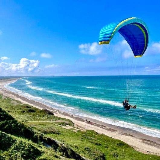 En person i sele under en blå‑grøn paraglider svæver over havet og bølgerne ved en lang, buet sandstrand omgivet af grønne klitter under en klar blå himmel.