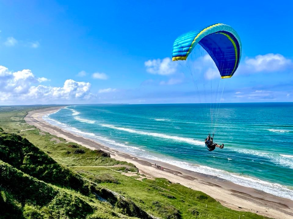 En person i sele under en blå‑grøn paraglider svæver over havet og bølgerne ved en lang, buet sandstrand omgivet af grønne klitter under en klar blå himmel.