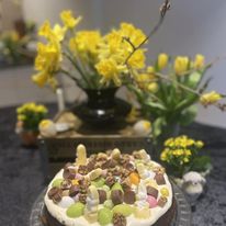 En rund kage i forgrunden dækket med hvid glasur og pyntet med små pastelfarvede chokoladeæg og hakket chokolade/nødder. Bagved står flere vaser med gule forårsblomster på et bord med mørk dug, hvilket giver et hyggeligt påskestemning.