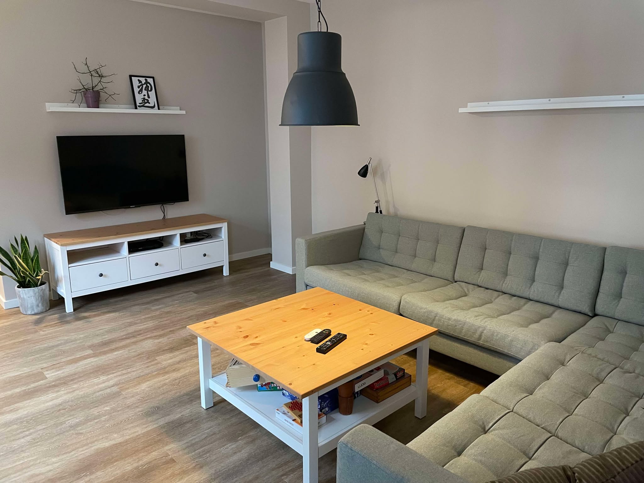 L-formet grå polstret sofa omkring et lyst træ-sofabord med to fjernbetjeninger, og et vægmonteret fladskærms-TV på en hvid mediebænk med en grøn potteplante ved siden af. En hvid væghylde over TV’et viser en indrammet sort kalligrafi med teksten u0022神武u0022.