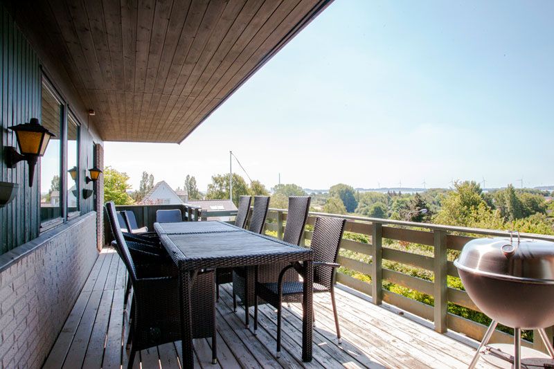 Træterrasse med et sort flettet havemøbelsæt bestående af et rektangulært bord og seks høje stole, arrangeret til udendørs spisning under et stort træudhæng med væglamper langs husvæggen. Til højre står en rund kuglegrill ved rækværket, og der er udsigt over grønne træer og tage i det fjerne.