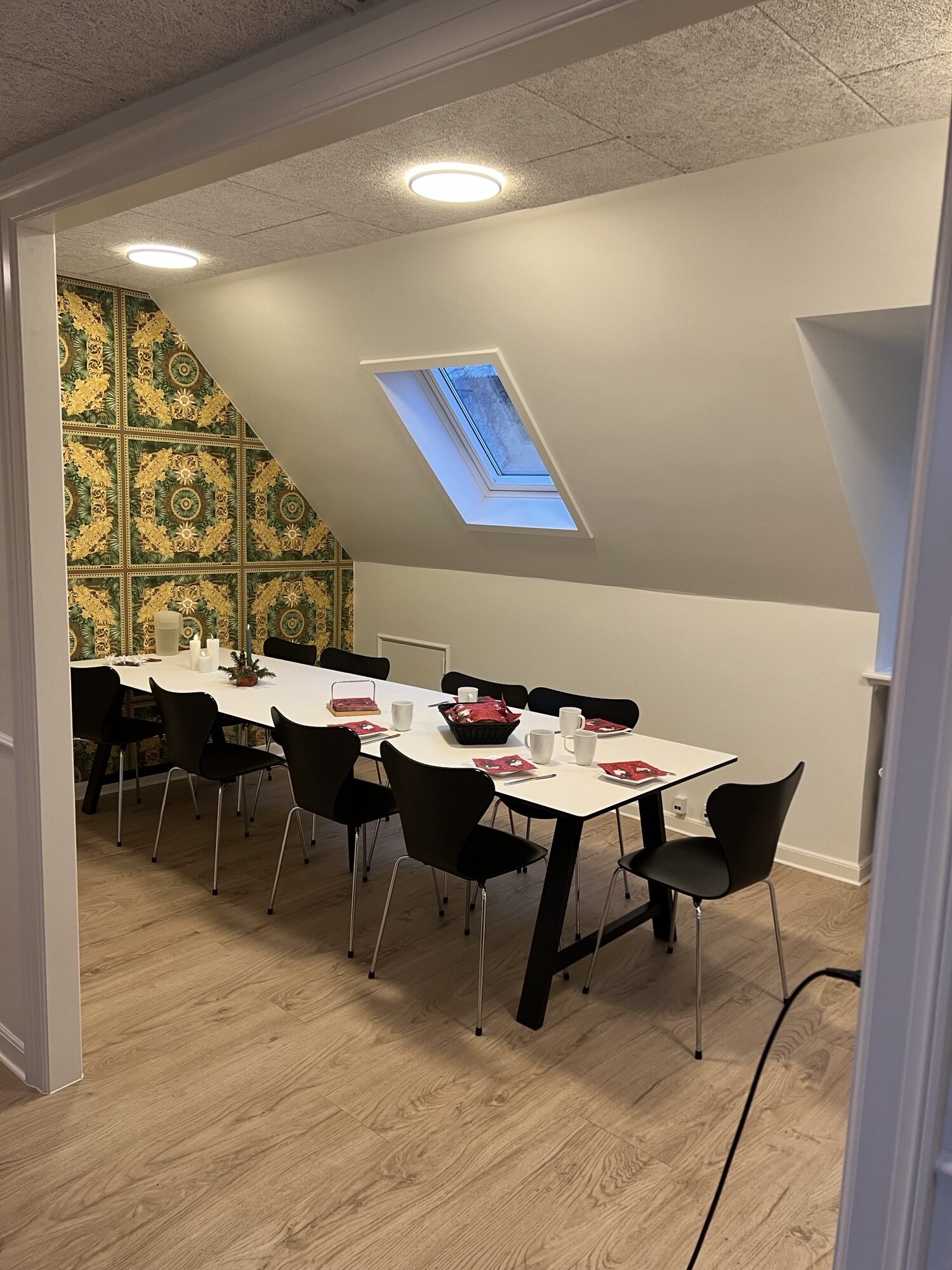 Langt hvidt bord dækket til kaffe med sorte stole omkring, hvide krus, røde servietter og skåle i midten af rummet. Rummet ligger under et skråt loft med tagvindue og har grøn-gult ornamenteret tapet på den ene væg og lyst trægulv.
