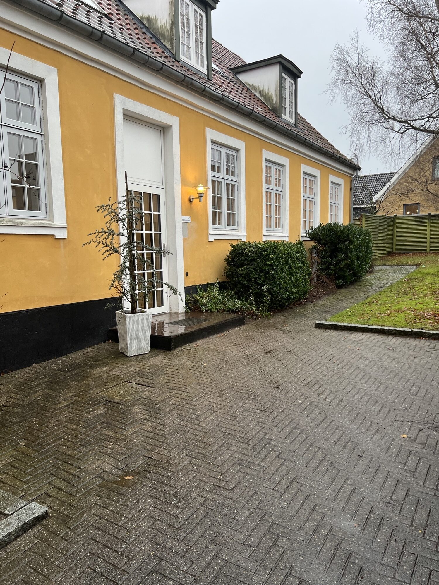 Gult hus med hvide vinduer og en hvid glasdør, en oplyst væglampe ved indgangen og en pottet plante på trinet; forpladsen er våd brostensbelægning i fiskebensmønster med buske langs facaden og en græsplet til højre under en grå himmel.