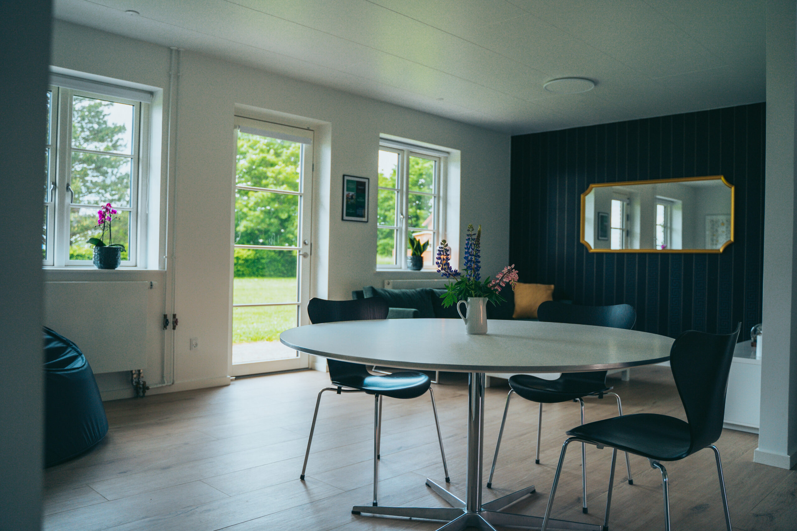 Rundt hvidt spisebord med en hvid kande fyldt med lupiner og vilde blomster står midt i rummet og er omgivet af fire sorte stole. Lyst opholdsrum med store vinduer og en glasdør ud til en grøn græsplæne, mørk træpanelvæg med et gyldent kantet spejl og en orkidé på vindueskarmen.