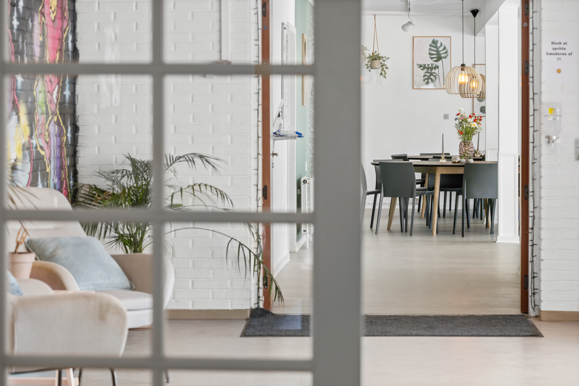 Gennem en rudemalet dør ses en lys stue og spisestue; til venstre en sofa med puder og grønne potteplanter, for enden et træbord med seks mørke stole, buket blomster, pendellampe og indrammet bladplakat. Skilt på højre væg med teksten: u0022Husk at spritte hænderne af !!!u0022.