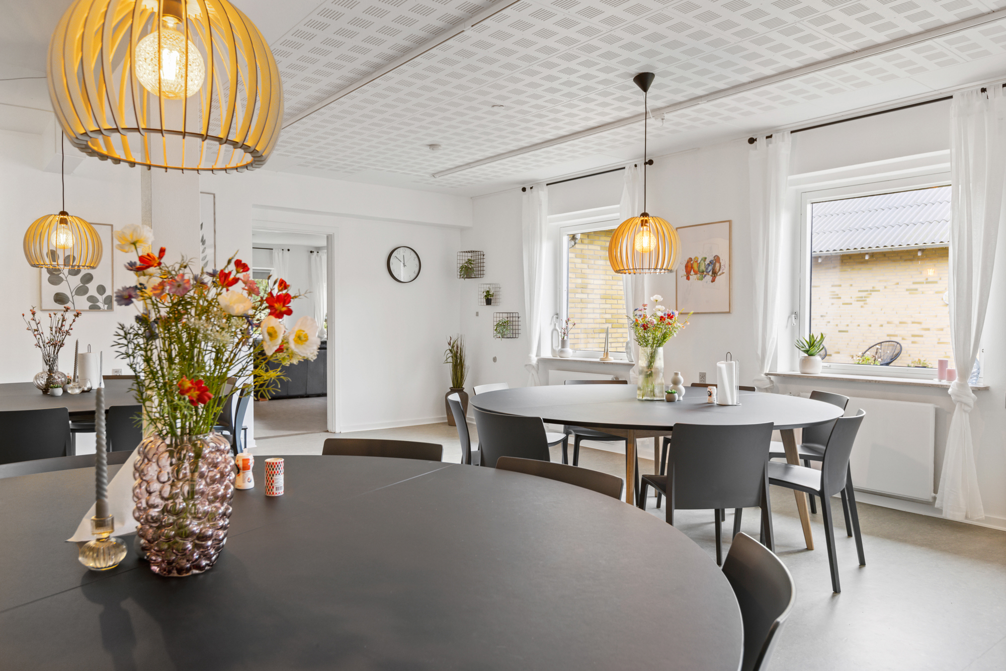 Rundt sort bord i forgrunden med en bobleglasvase fyldt med farverige blomster; i baggrunden flere runde spiseborde med grå stole, hængende træpendler, hvide gardiner og et vægur på en lys væg i et lyst fællesrum.