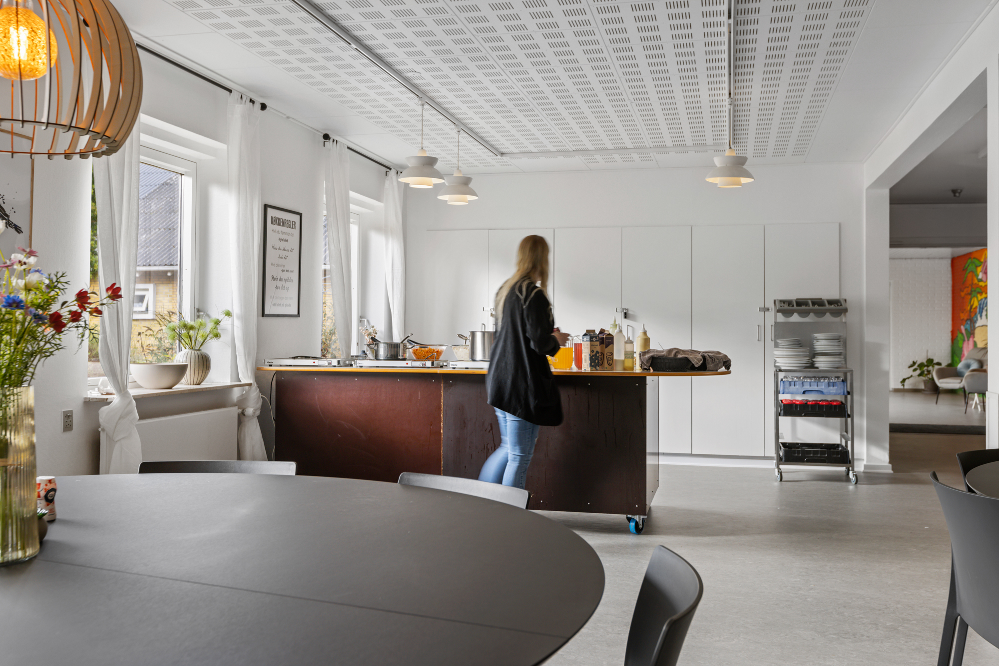 Lyse fælleskøkken med et rundt bord i forgrunden og en kvinde, der står ved en mørk køkkenø og rækker efter flasker og glas. Store vinduer med hvide gardiner, hængelamper og en rullevogn med tallerkener ses til højre, og gennem døren skimtes et opholdsområde med farverigt vægmaleri.