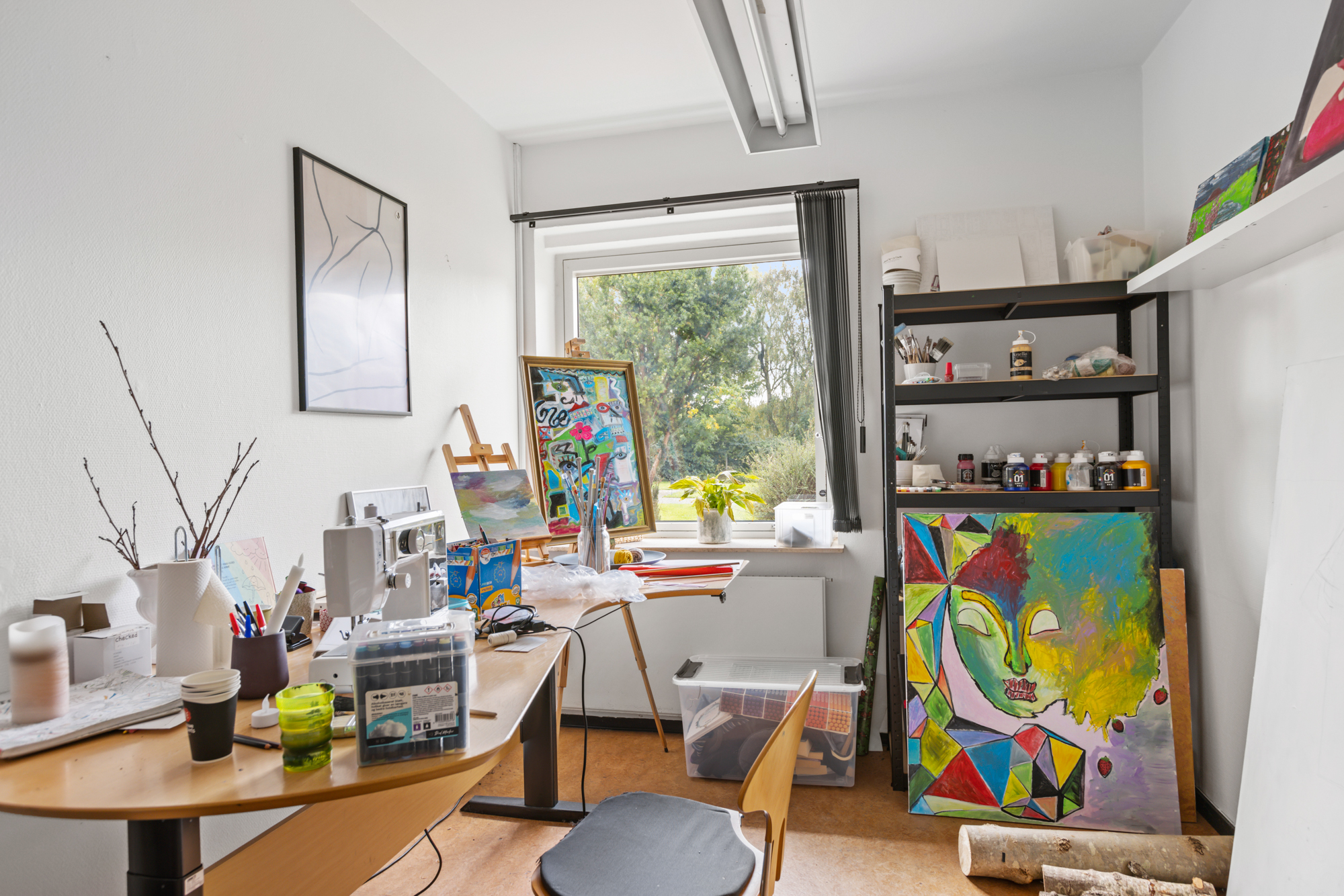Lyst malerværksted med et buet arbejdsbord dækket af pensler, krus, en symaskine og plastikbokse foran et vindue med udsigt til træer. Et staffeli med farverig collage står ved vinduet, og en reol med malertuber samt et stort geometrisk, farverigt portrætmaleri lænet mod væggen.