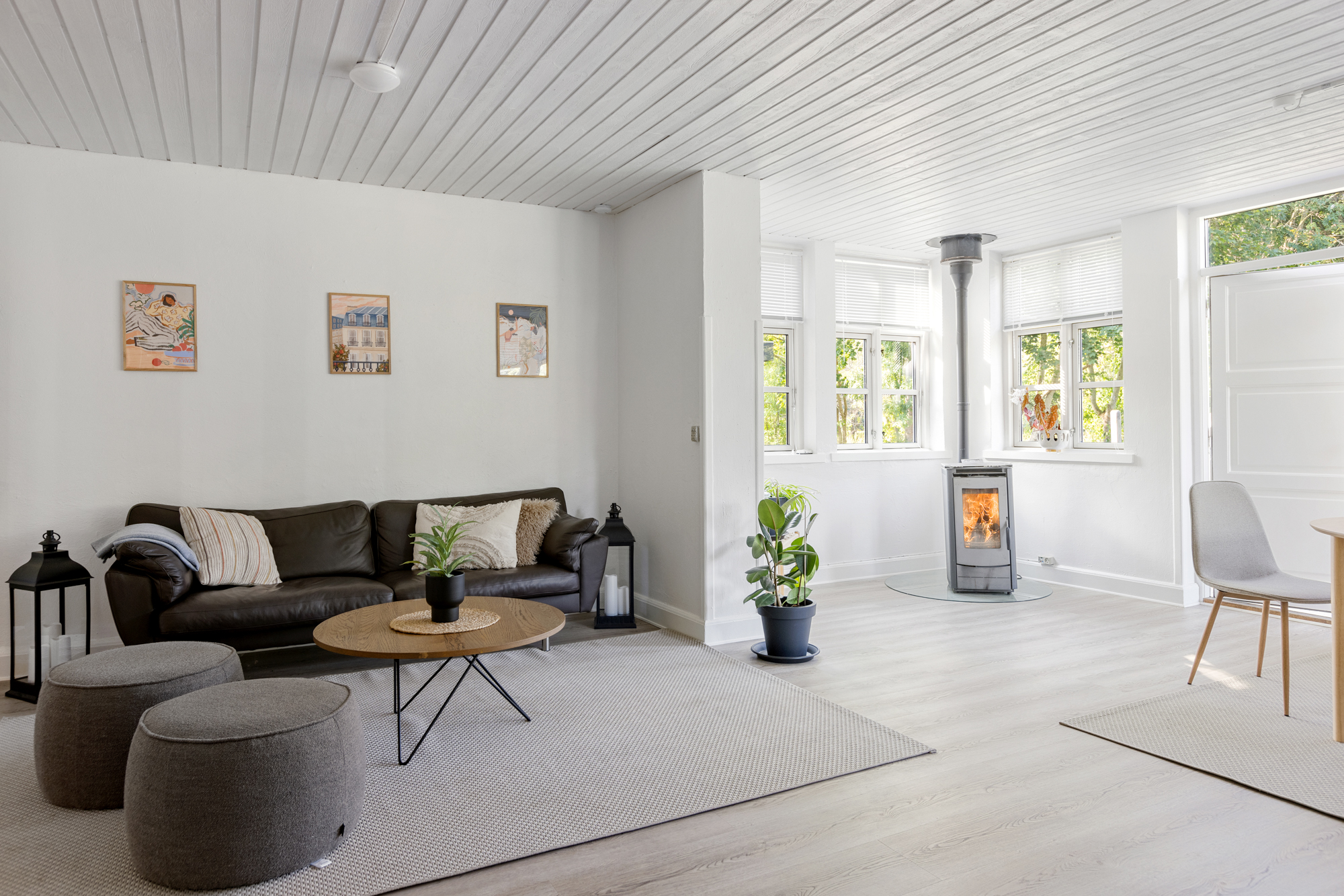 Stue med en mørk lædersofa og et rundt sofabord på et lyst tæppe, flankeret af to grå puffer og to store lanterner; tre små indrammede billeder hænger på den hvide væg over sofaen. I baggrunden ses en tændt brændeovn foran vinduer, en grøn potteplante på gulvet og en enkel spiseplads til højre i et lyst, minimalistisk rum.